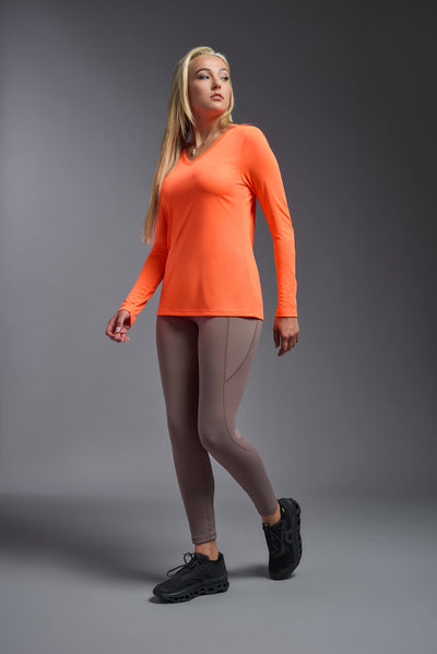 A female model with short brown hair on a gray background, wearing the Women's DriMax™ V-neck Long Sleeve Expert Tee in the color safety orange from a full body side view. This shirt is made in the USA and is quick-drying and moisture-wicking. #color_safety-orange