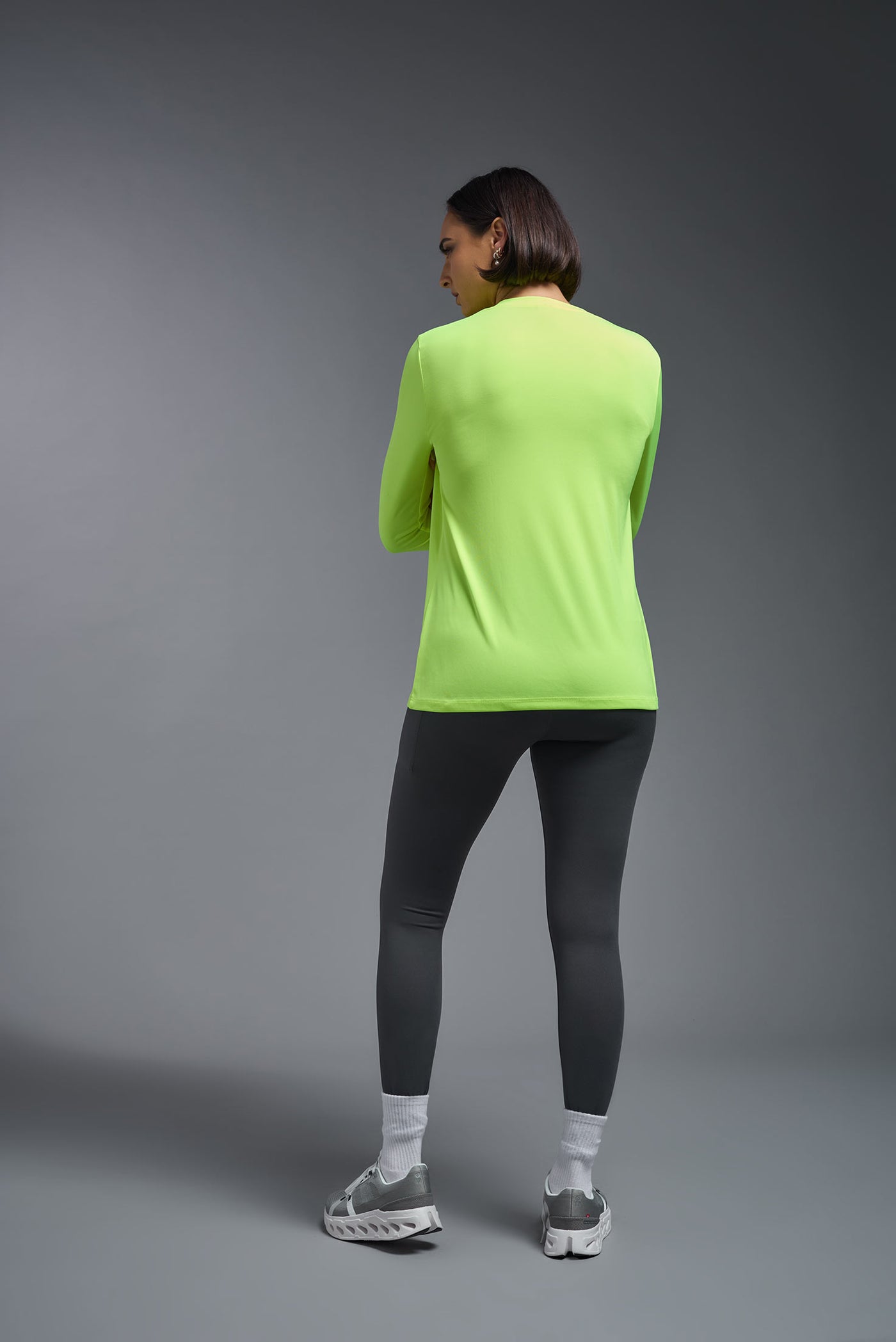 A female model with short brown hair on a gray background, wearing the Women's DriMax™ V-neck Long Sleeve Expert Tee in the color safety yellow from a full body back view. This shirt is made in the USA and is quick-drying and moisture-wicking. #color_safety-yellow