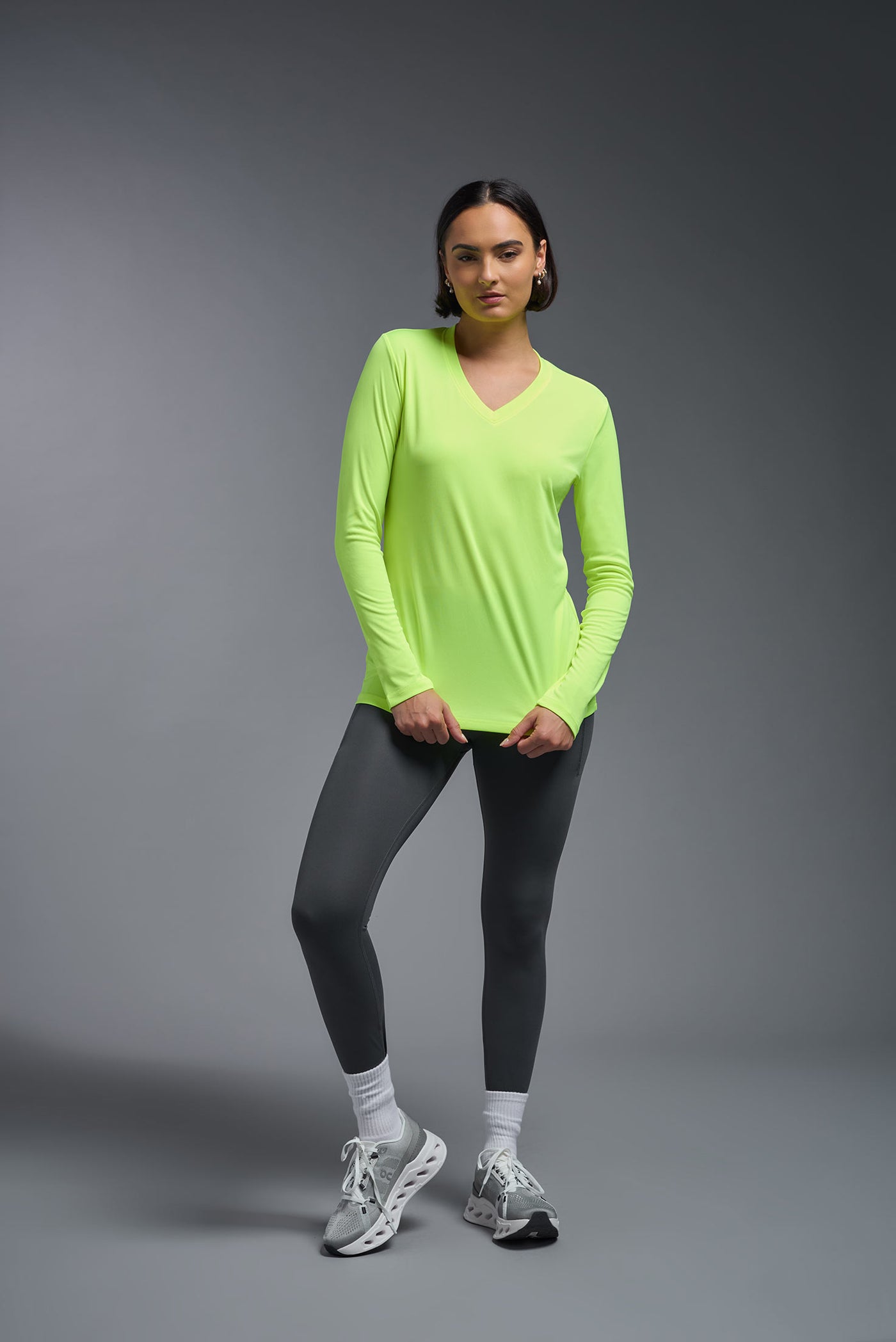 A female model with short brown hair on a gray background, wearing the Women's DriMax™ V-neck Long Sleeve Expert Tee in the color safety yellow from a full body front view. This shirt is made in the USA and is quick-drying and moisture-wicking. #color_safety-yellow