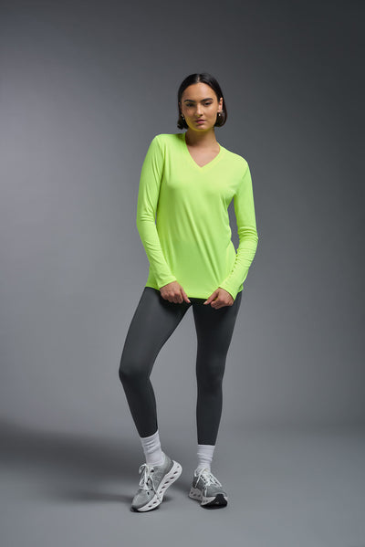 A female model with short brown hair on a gray background, wearing the Women's DriMax™ V-neck Long Sleeve Expert Tee in the color safety yellow from a full body front view. This shirt is made in the USA and is quick-drying and moisture-wicking. #color_safety-yellow