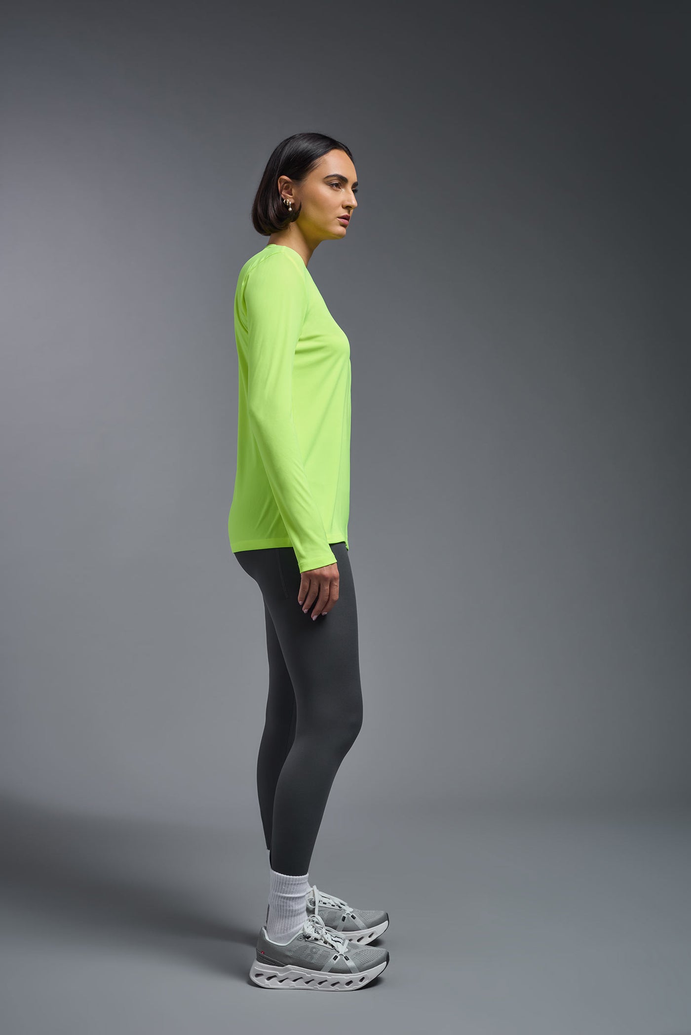 A female model with short brown hair on a gray background, wearing the Women's DriMax™ V-neck Long Sleeve Expert Tee in the color safety yellow from a full body side view. This shirt is made in the USA and is quick-drying and moisture-wicking. #color_safety-yellow