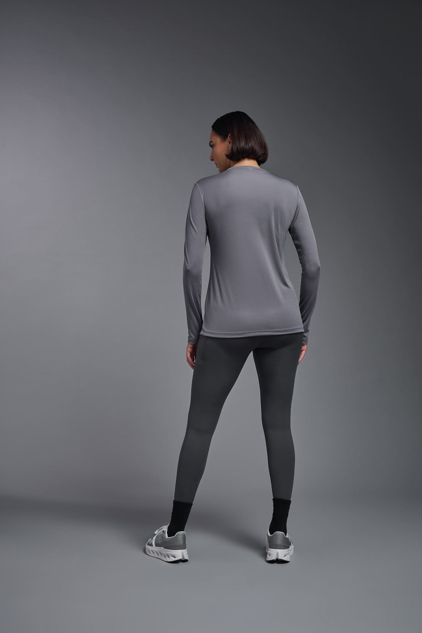A female model with short brown hair on a gray background, wearing the Women's DriMax™ V-neck Long Sleeve Expert Tee in the color steel gray from a full body back view. This shirt is made in the USA and is quick-drying and moisture-wicking. #color_steel