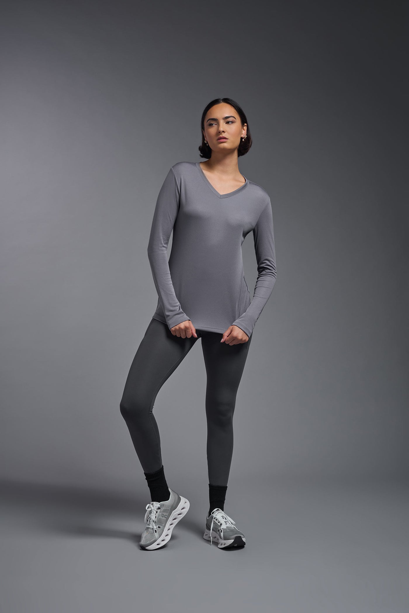 A female model with short brown hair on a gray background, wearing the Women's DriMax™ V-neck Long Sleeve Expert Tee in the color steel gray from a full body front view. This shirt is made in the USA and is quick-drying and moisture-wicking. #color_steel