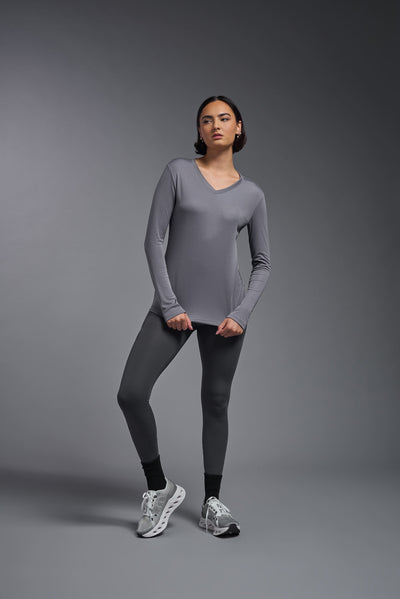 A female model with short brown hair on a gray background, wearing the Women's DriMax™ V-neck Long Sleeve Expert Tee in the color steel gray from a full body front view. This shirt is made in the USA and is quick-drying and moisture-wicking. #color_steel