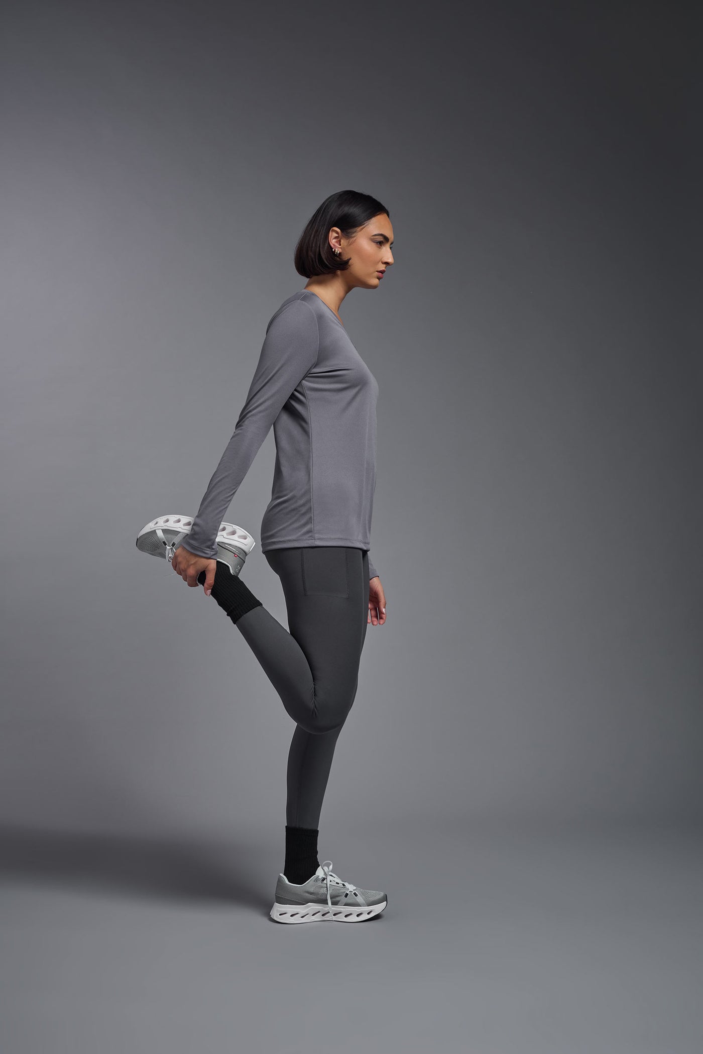 A female model with short brown hair on a gray background, wearing the Women's DriMax™ V-neck Long Sleeve Expert Tee in the color steel gray from a full body side view. This shirt is made in the USA and is quick-drying and moisture-wicking. #color_steel