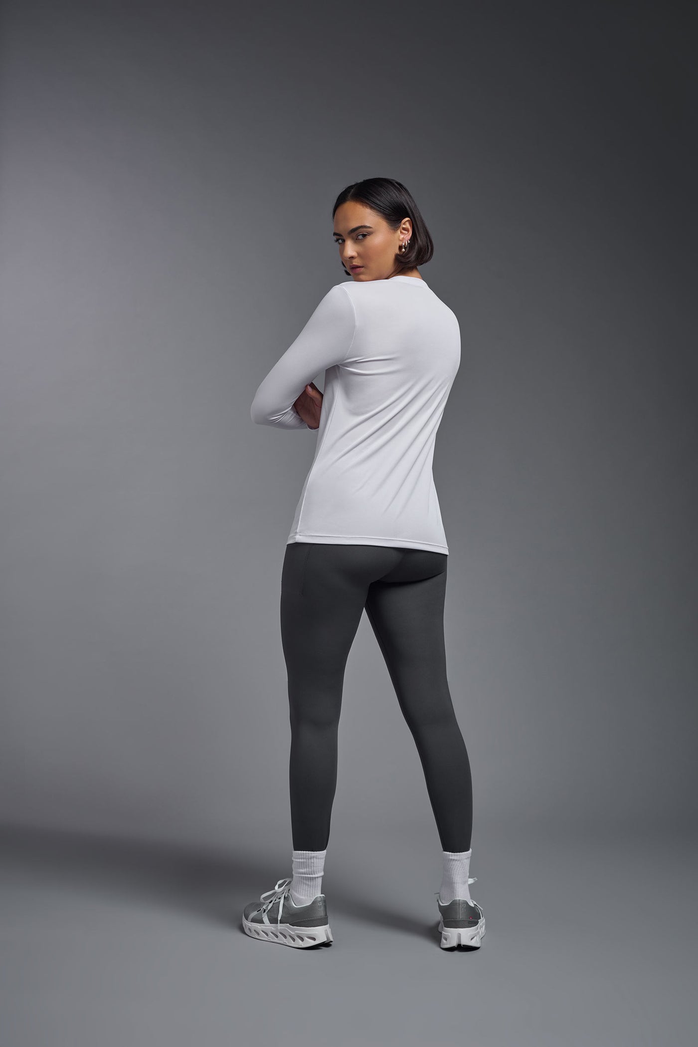 A female model with short brown hair on a gray background, wearing the Women's DriMax™ V-neck Long Sleeve Expert Tee in the color white from a full body back view. This shirt is made in the USA and is quick-drying and moisture-wicking. #color_white