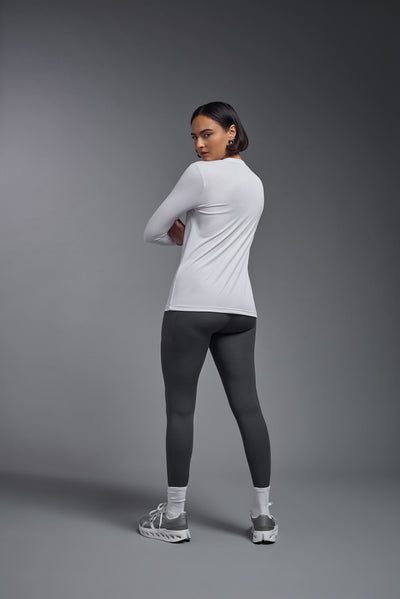 A female model with short brown hair on a gray background, wearing the Women's DriMax™ V-neck Long Sleeve Expert Tee in the color white from a full body back view. This shirt is made in the USA and is quick-drying and moisture-wicking. #color_white
