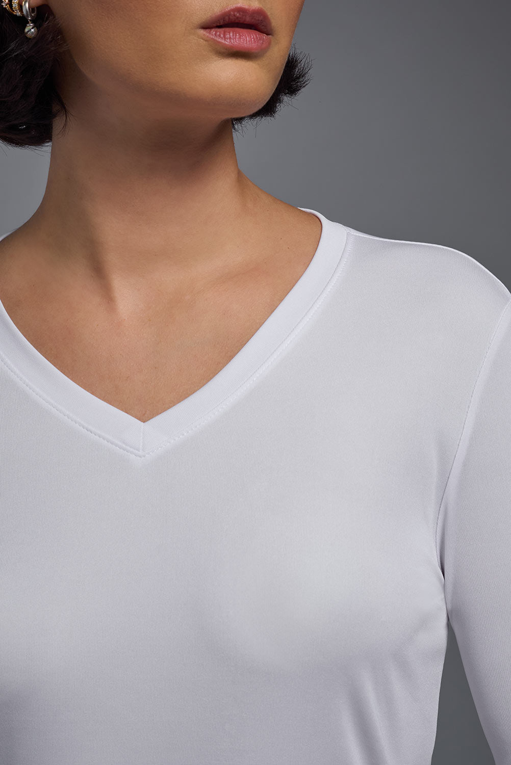 A female model with short brown hair on a gray background, wearing the Women's DriMax™ V-neck Long Sleeve Expert Tee in the color white from a closeup collar front view. This shirt is made in the USA and is quick-drying and moisture-wicking. #color_white