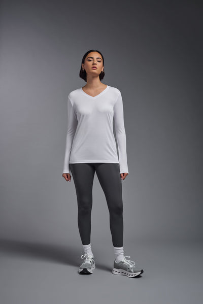 A female model with short brown hair on a gray background, wearing the Women's DriMax™ V-neck Long Sleeve Expert Tee in the color white from a full body front view. This shirt is made in the USA and is quick-drying and moisture-wicking. #color_white