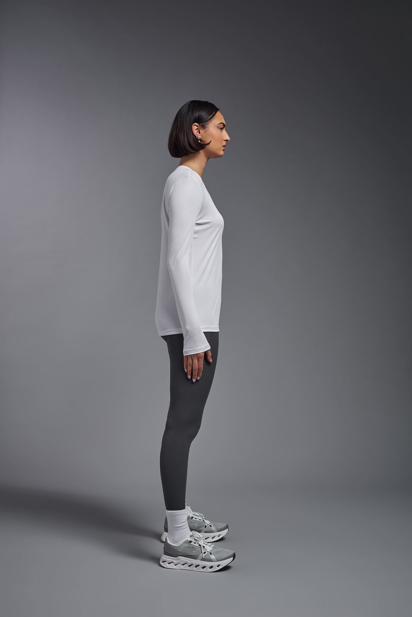 A female model with short brown hair on a gray background, wearing the Women's DriMax™ V-neck Long Sleeve Expert Tee in the color white from a full body side view. This shirt is made in the USA and is quick-drying and moisture-wicking. #color_white