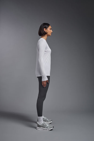 A female model with short brown hair on a gray background, wearing the Women's DriMax™ V-neck Long Sleeve Expert Tee in the color white from a full body side view. This shirt is made in the USA and is quick-drying and moisture-wicking. #color_white