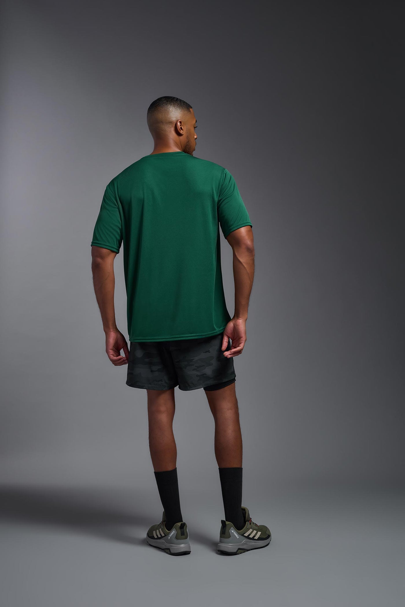 An African American male model with a beard on a gray background, wearing the Men's DriMax Expert Tee Shirt in the color forest green from a full body back view. This shirt is made in the USA and is quick-drying and moisture-wicking. #color_forest-green