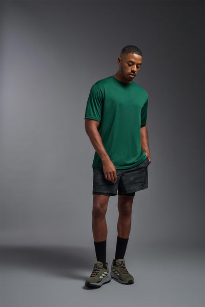 An African American male model with a beard on a gray background, wearing the Men's DriMax Expert Tee Shirt in the color forest green from a full body view. This shirt is made in the USA and is quick-drying and moisture-wicking. #color_forest-green