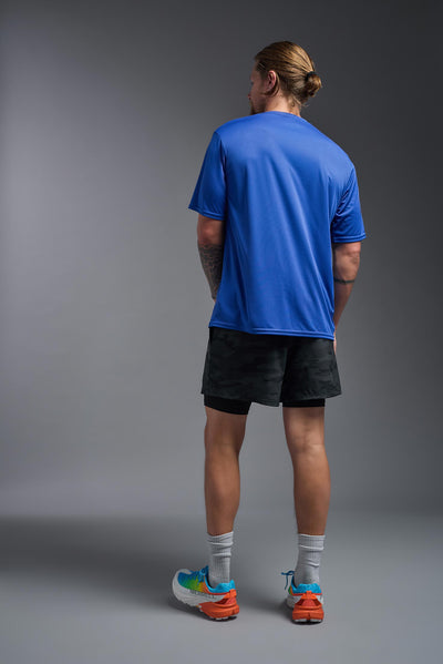 A blonde male model with a beard on a gray background, wearing the Men's DriMax Expert Tee Shirt in the color royal blue from a full body back view. This shirt is made in the USA and is quick-drying and moisture-wicking. #color_royal-blue