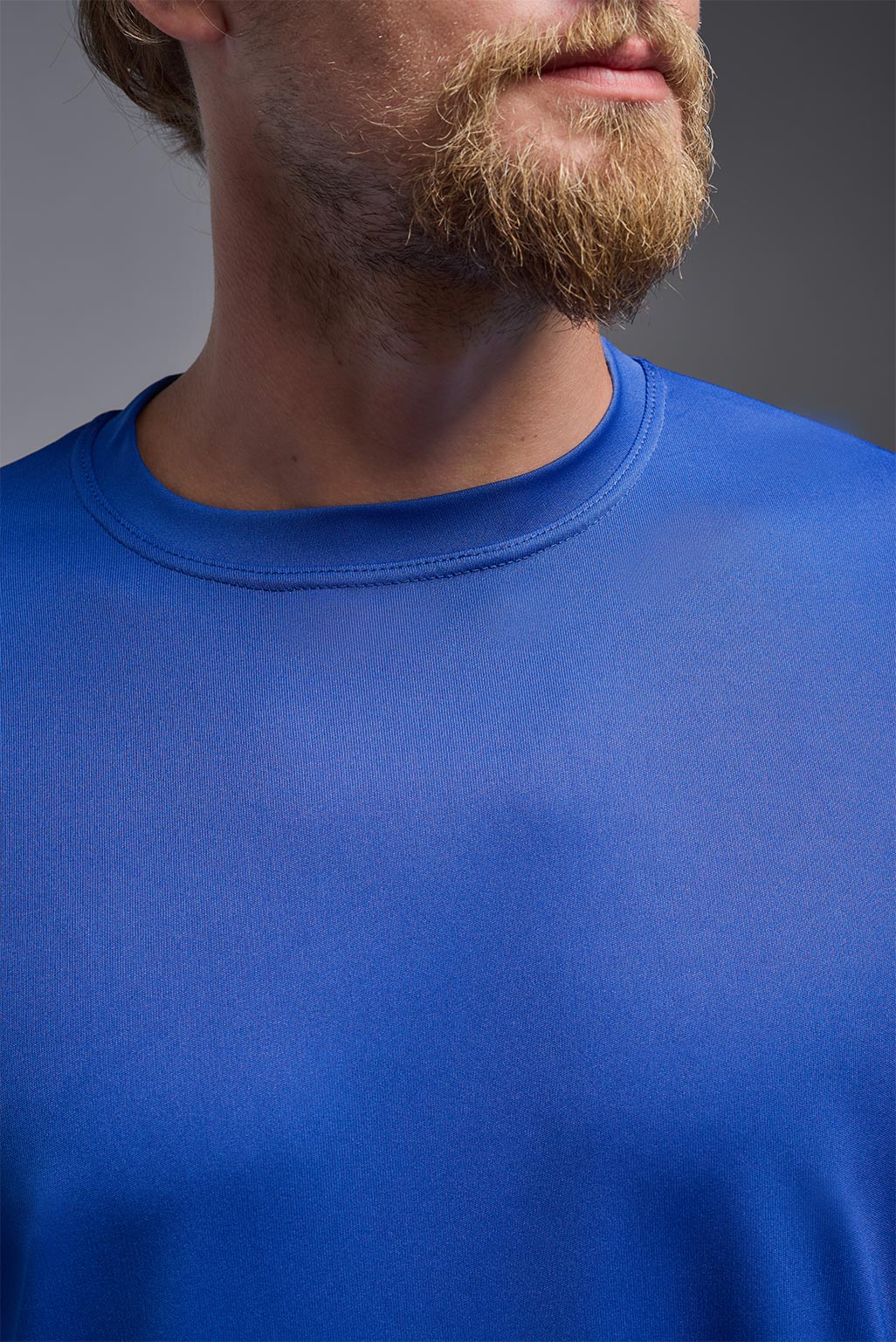A blonde male model with a beard on a gray background, wearing the Men's DriMax Expert Tee Shirt in the color royal blue from a close-up collar view. This shirt is made in the USA and is quick-drying and moisture-wicking. #color_royal-blue