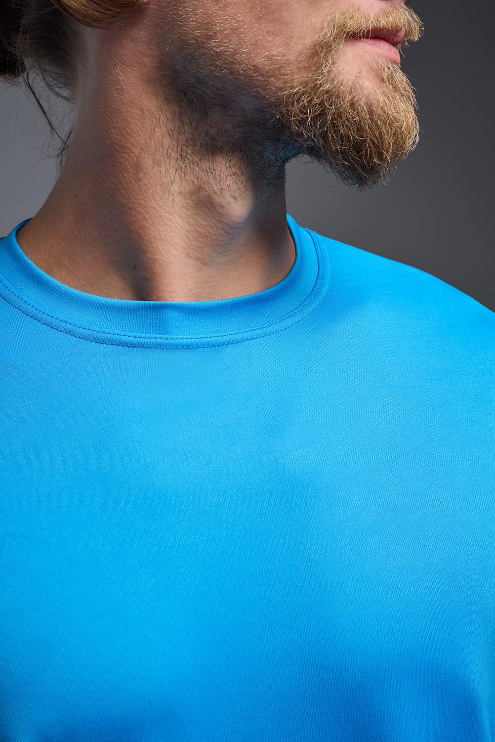 A blonde male model with a beard on a gray background, wearing the Men's DriMax shirt in the color safety blue from a front close-up collar view. This shirt is made in the USA and is quick-drying and moisture-wicking. #color_safety-blue