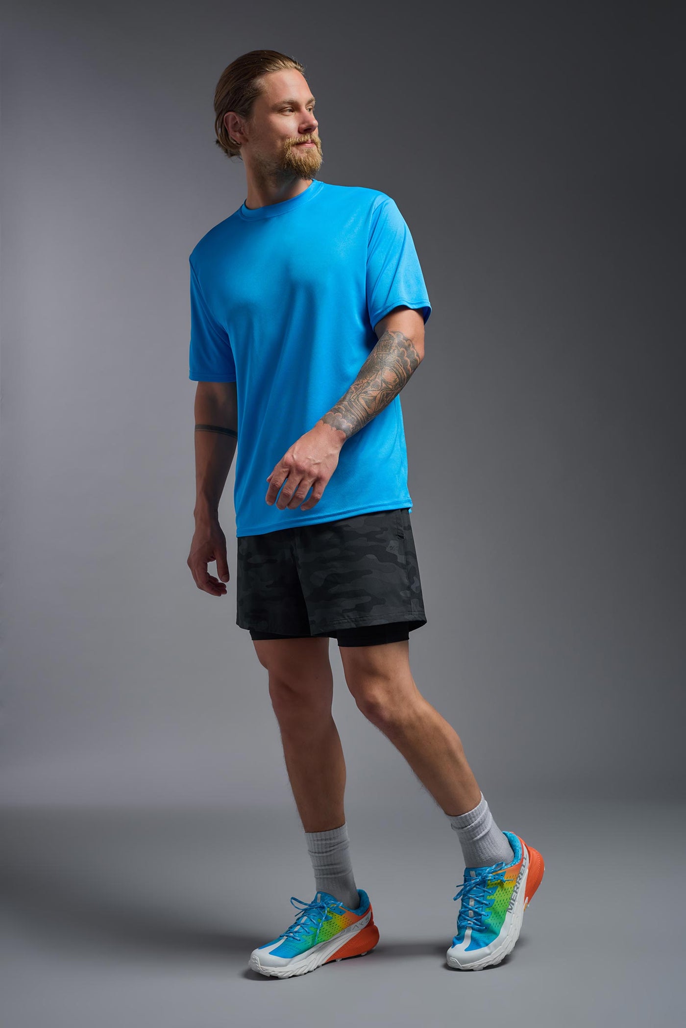 A blonde male model with a beard on a gray background, wearing the Men's DriMax shirt in the color safety blue from a full body side view. This shirt is made in the USA and is quick-drying and moisture-wicking. #color_safety-blue