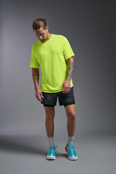 A blonde male model with a beard on a gray background, wearing the Men's DriMax shirt in the color safety yellow from a full body front view. This shirt is made in the USA and is quick-drying and moisture-wicking. #color_safety-yellow