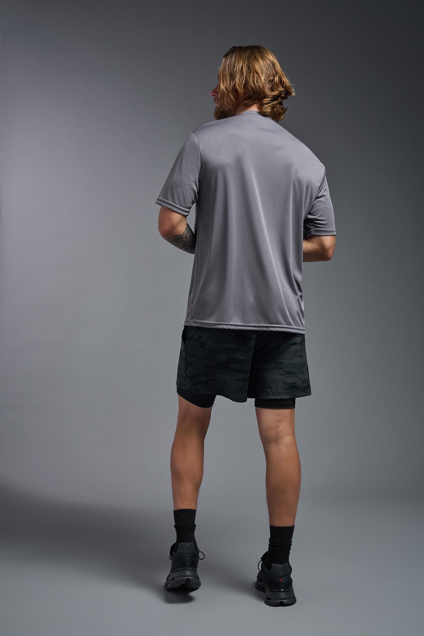 A blonde male model with a beard on a gray background, wearing the Men's DriMax Expert Tee Shirt in the color steel from a full body back view. This shirt is made in the USA and is quick-drying and moisture-wicking. #color_steel
