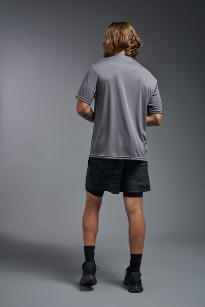 A blonde male model with a beard on a gray background, wearing the Men's DriMax Expert Tee Shirt in the color steel from a full body back view. This shirt is made in the USA and is quick-drying and moisture-wicking. #color_steel