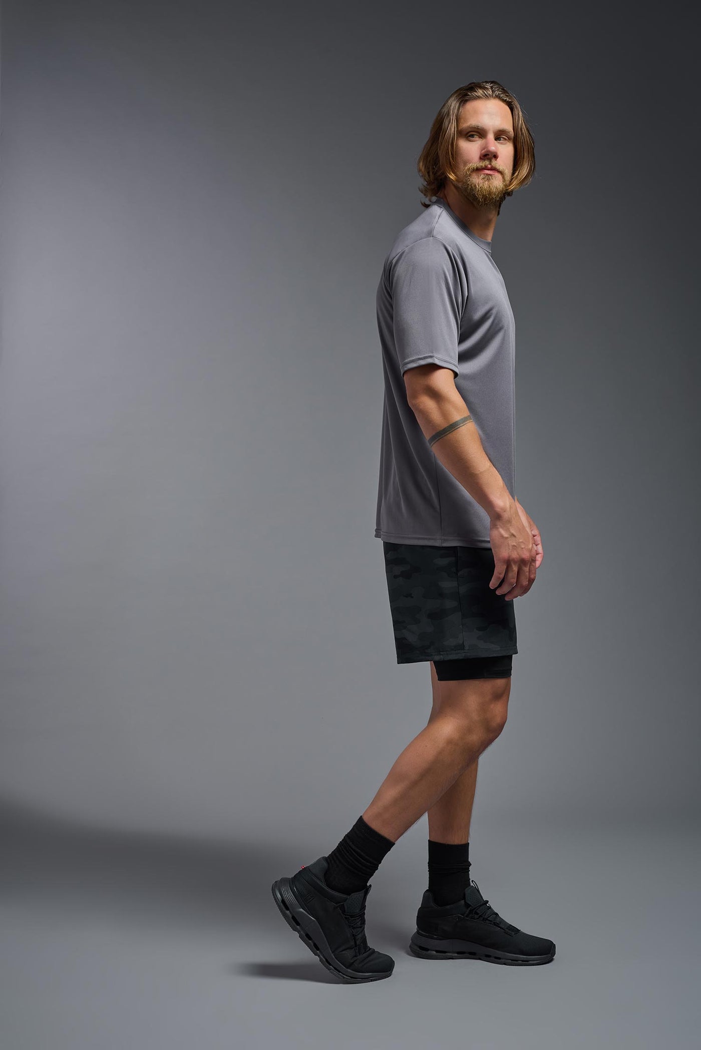 A blonde male model with a beard on a gray background, wearing the Men's DriMax Expert Tee Shirt in the color steel from a full body side view. This shirt is made in the USA and is quick-drying and moisture-wicking. #color_steel