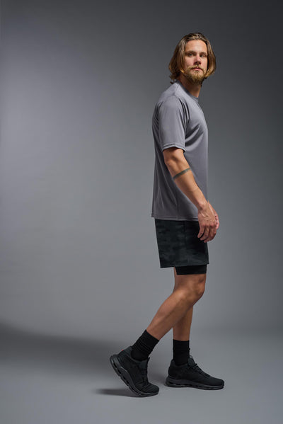 A blonde male model with a beard on a gray background, wearing the Men's DriMax Expert Tee Shirt in the color steel from a full body side view. This shirt is made in the USA and is quick-drying and moisture-wicking. #color_steel