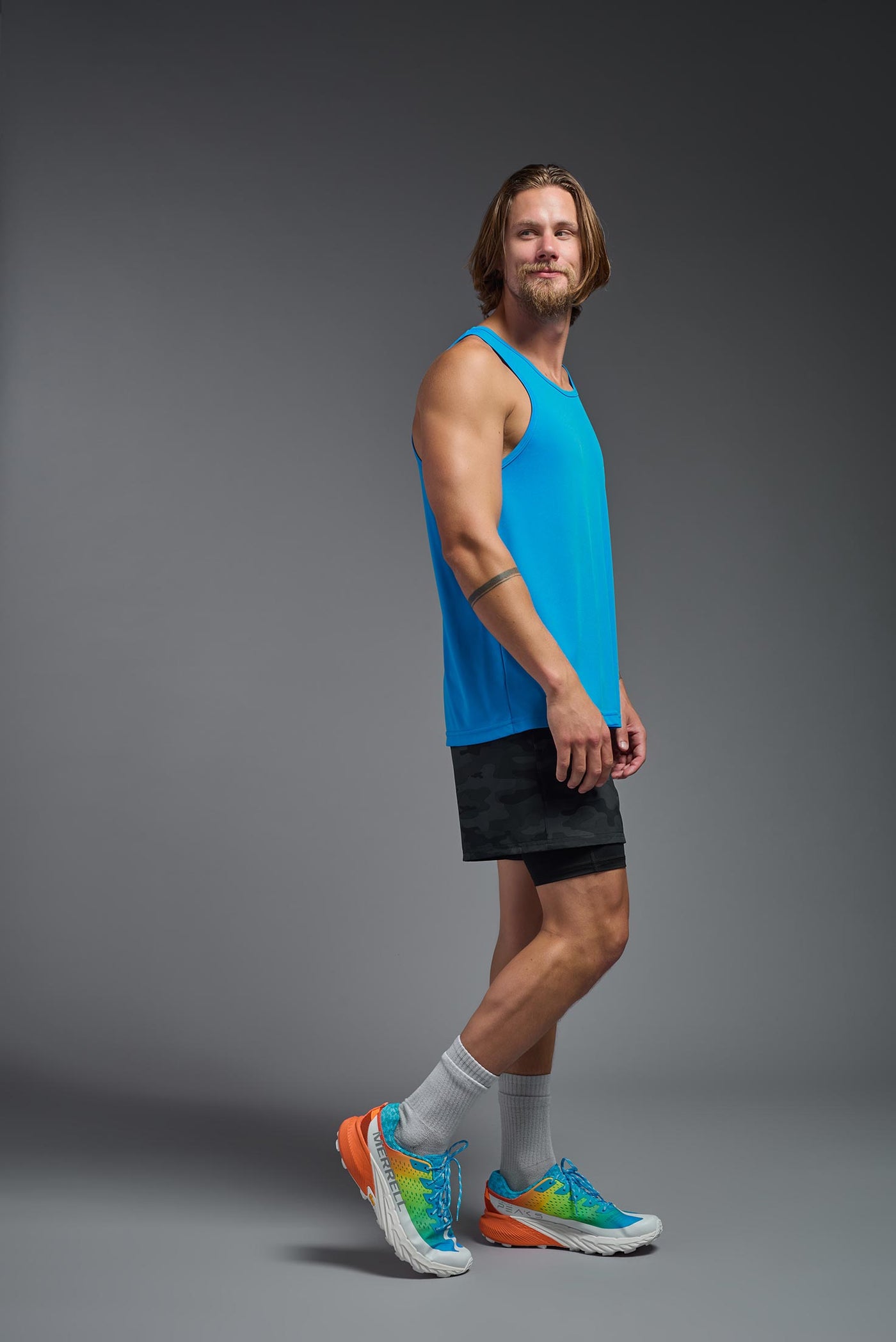 A blonde male model with a beard on a gray background, wearing the Men's DriMax Endurance Tank in the color safety blue from a full body side view. This shirt is made in the USA and is quick-drying and moisture-wicking. #color_safety-blue