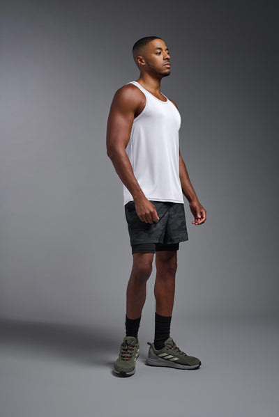 A blonde male model with a beard on a gray background, wearing the Men's DriMax Endurance Tank in the color white from a full body side view. This shirt is made in the USA and is quick-drying and moisture-wicking. #color_white