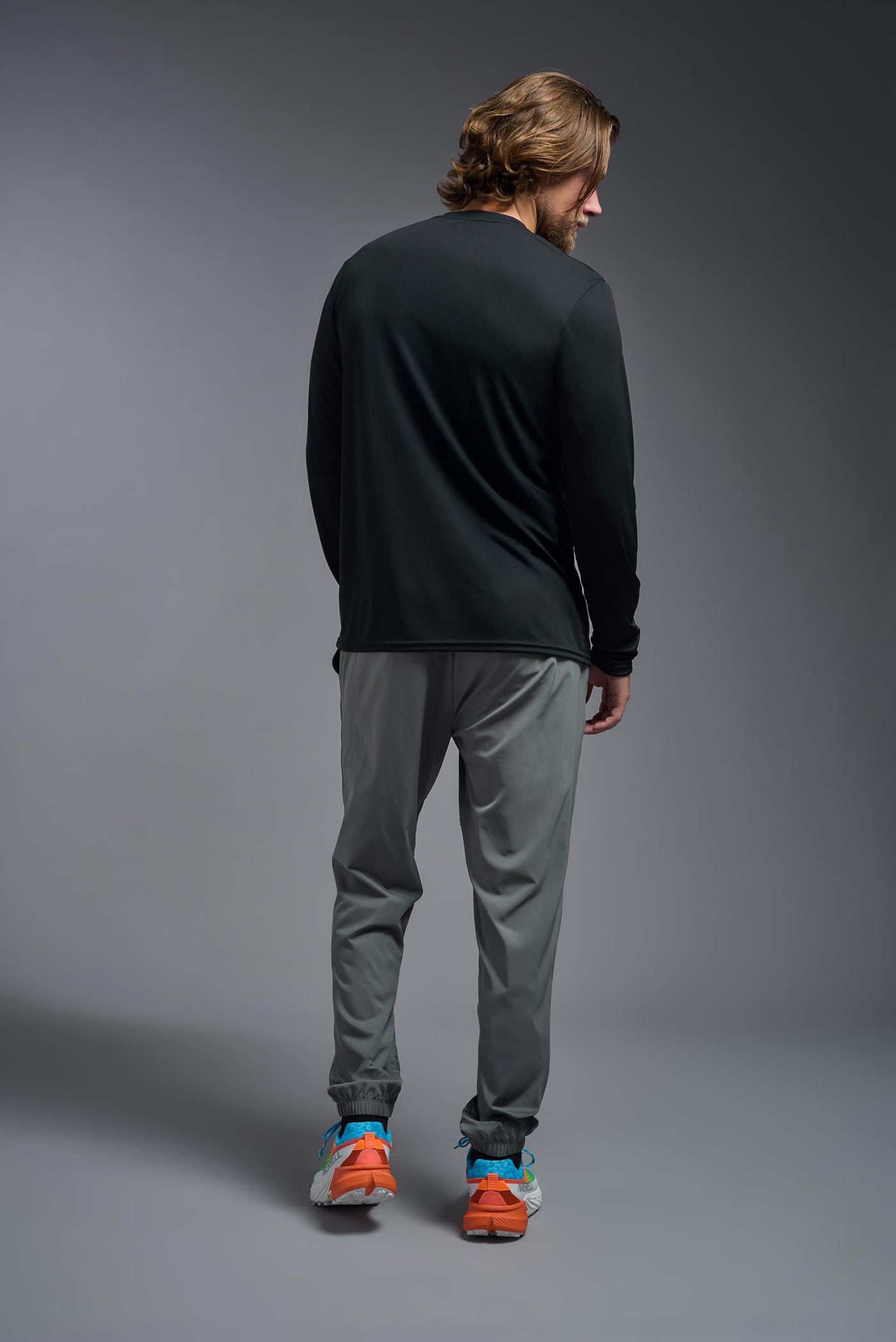 A blonde male model with a beard on a gray background, wearing the Men's DriMax Long Sleeve Expert Shirt in the color black from a full body back view. This shirt is made in the USA and is quick-drying and moisture-wicking. #color_black
