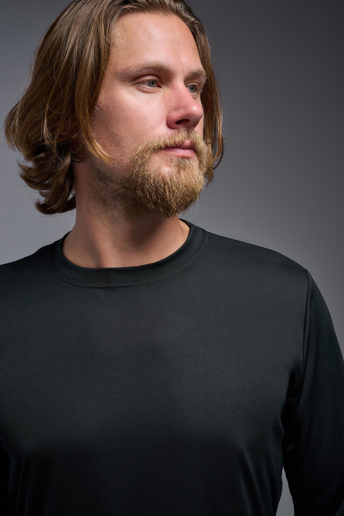 A blonde male model with a beard on a gray background, wearing the Men's DriMax Long Sleeve Expert Shirt in the color black from a close-up collar view. This shirt is made in the USA and is quick-drying and moisture-wicking. #color_black