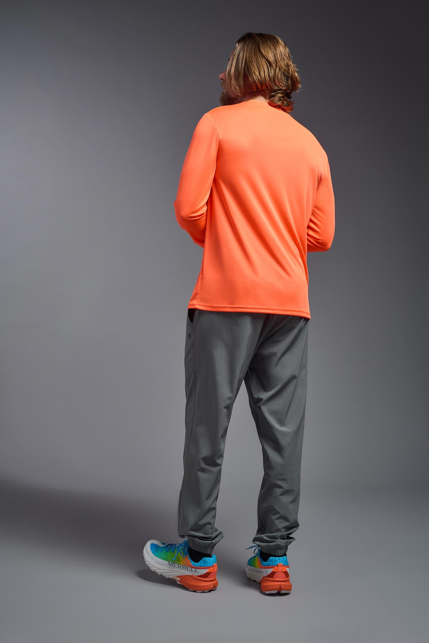 A blonde male model with a beard on a gray background, wearing the Men's DriMax Long Sleeve Expert Shirt in the color safety orange from a full body back view. This shirt is made in the USA and is quick-drying and moisture-wicking. #color_safety-orange