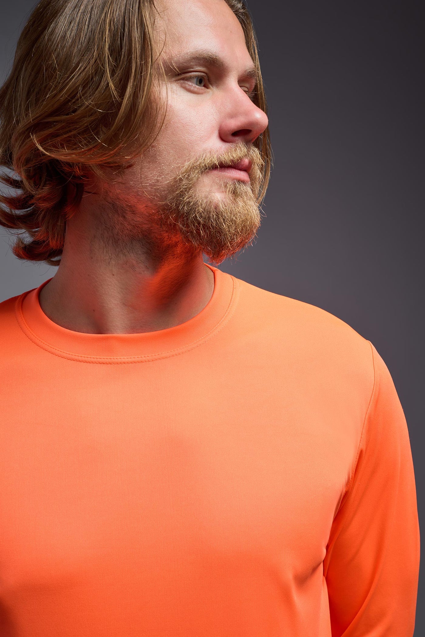 A blonde male model with a beard on a gray background, wearing the Men's DriMax Long Sleeve Expert shirt in the color safety orange from a full body close-up view. This shirt is made in the USA and is quick-drying and moisture-wicking. #color_safety-orange