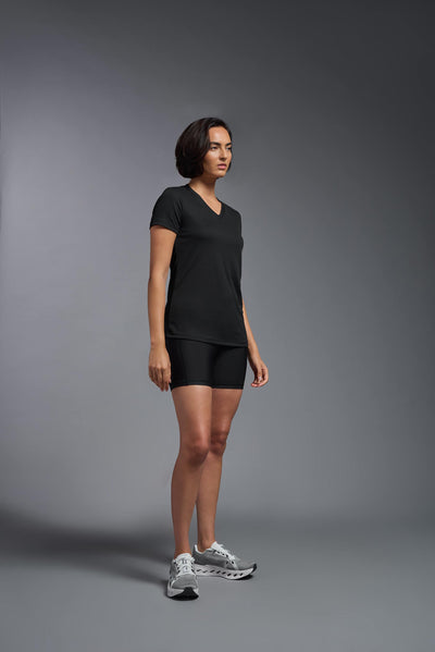 A female model with brown short hair on a gray background, wearing the Women's Oxymesh™ V-neck Tech Tee in the color black from a full body side view. This shirt is made in the USA and is quick-drying and moisture-wicking. #color_black