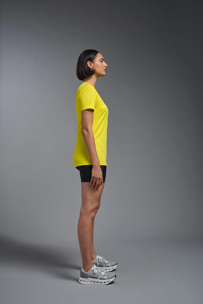 A female model with brown short hair on a gray background, wearing the Women's Oxymesh™ V-neck Tech Tee in the color bright yellow from a full body side view. This shirt is made in the USA and is quick-drying and moisture-wicking. #color_bright-yellow