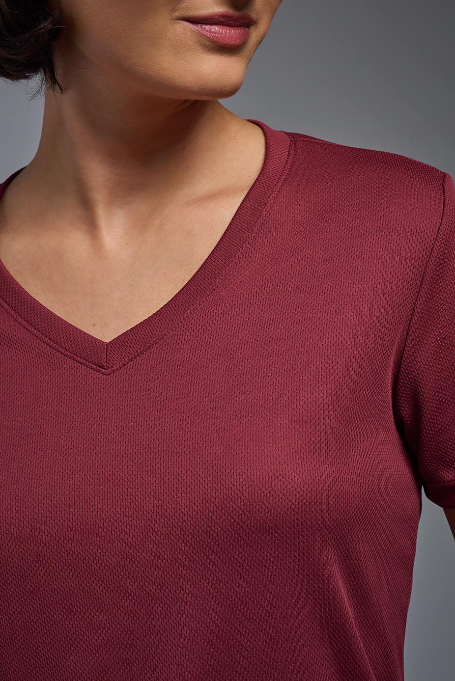 A female model with brown short hair on a gray background, wearing the Women's Oxymesh™ V-neck Tech Tee in the color cardinal from a closeup collar front view. This shirt is made in the USA and is quick-drying and moisture-wicking. #color_cardinal