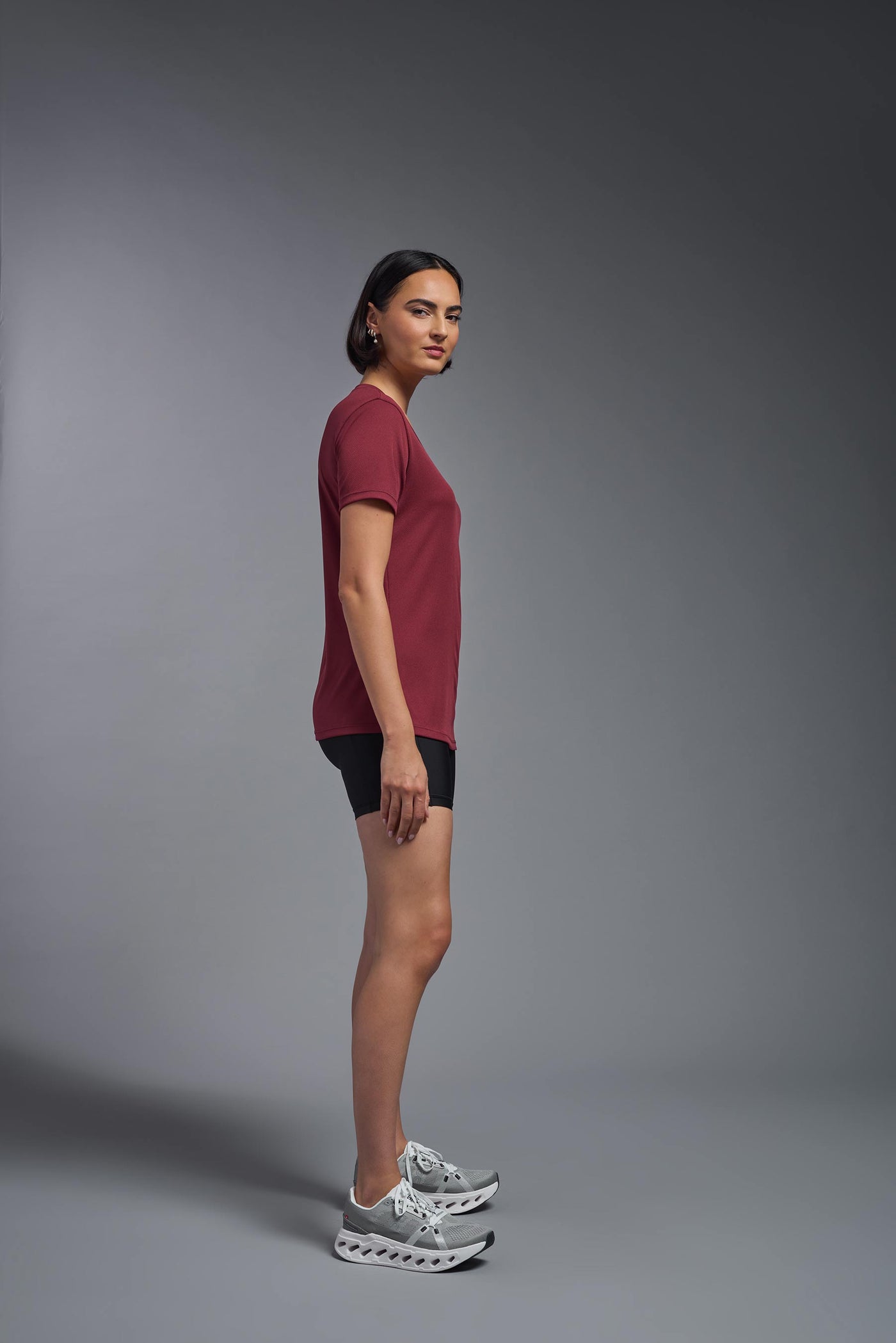 A female model with brown short hair on a gray background, wearing the Women's Oxymesh™ V-neck Tech Tee in the color cardinal from a full body side view. This shirt is made in the USA and is quick-drying and moisture-wicking. #color_cardinal