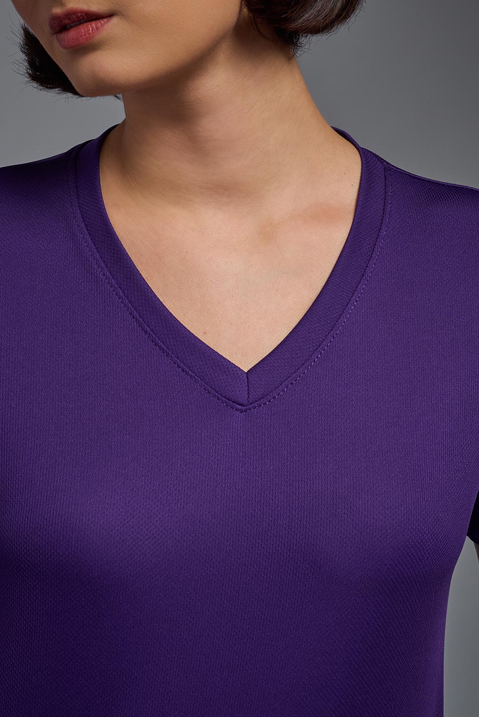 A female model with short brown hair on a gray background, wearing the Women's Oxymesh™ V-neck Tech Tee in the color dark purple from a closeup collar view. This shirt is made in the USA and is quick-drying and moisture-wicking. #color_dark-purple