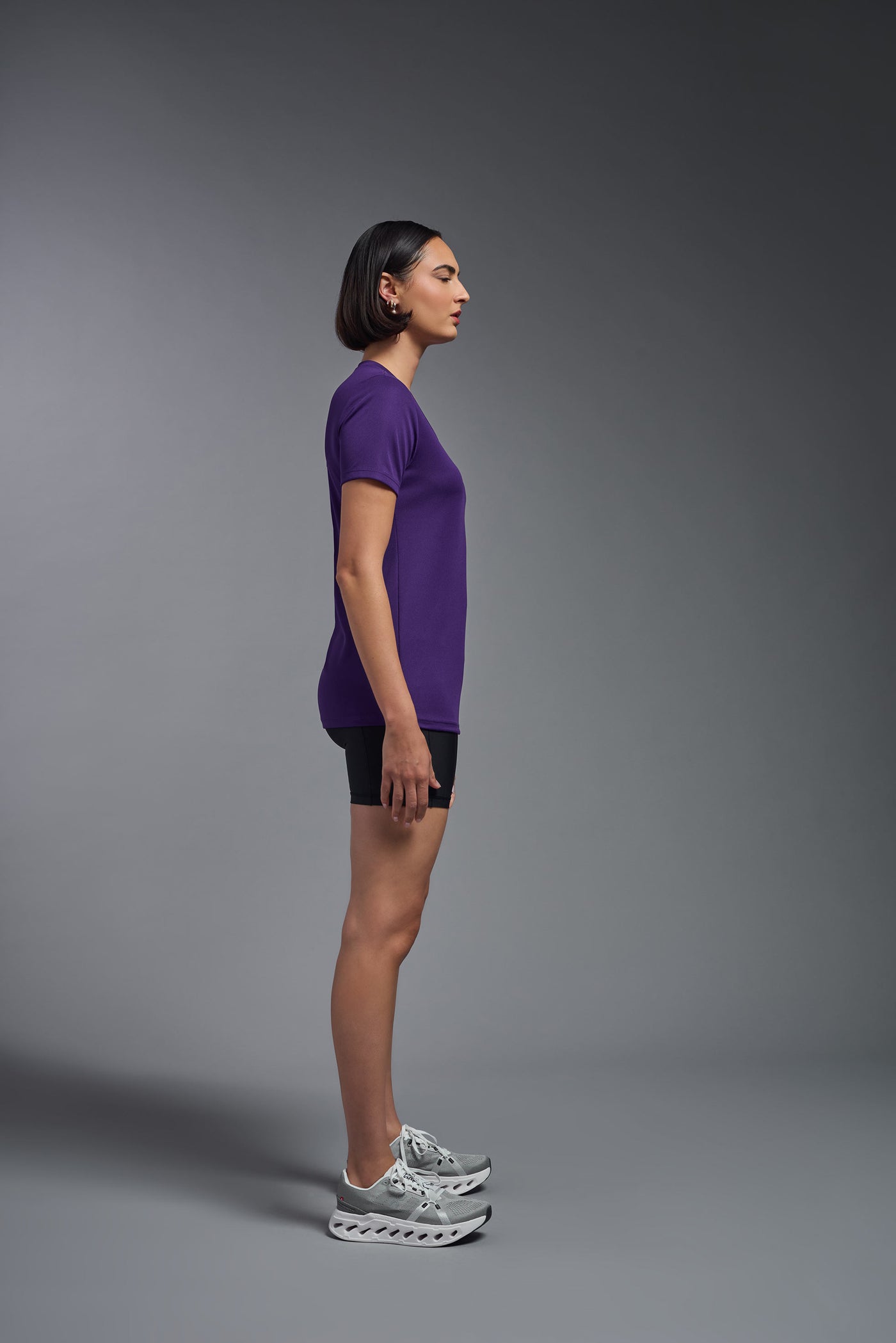 A female model with short brown hair on a gray background, wearing the Women's Oxymesh™ V-neck Tech Tee in the color dark purple from a full body side view. This shirt is made in the USA and is quick-drying and moisture-wicking. #color_dark-purple