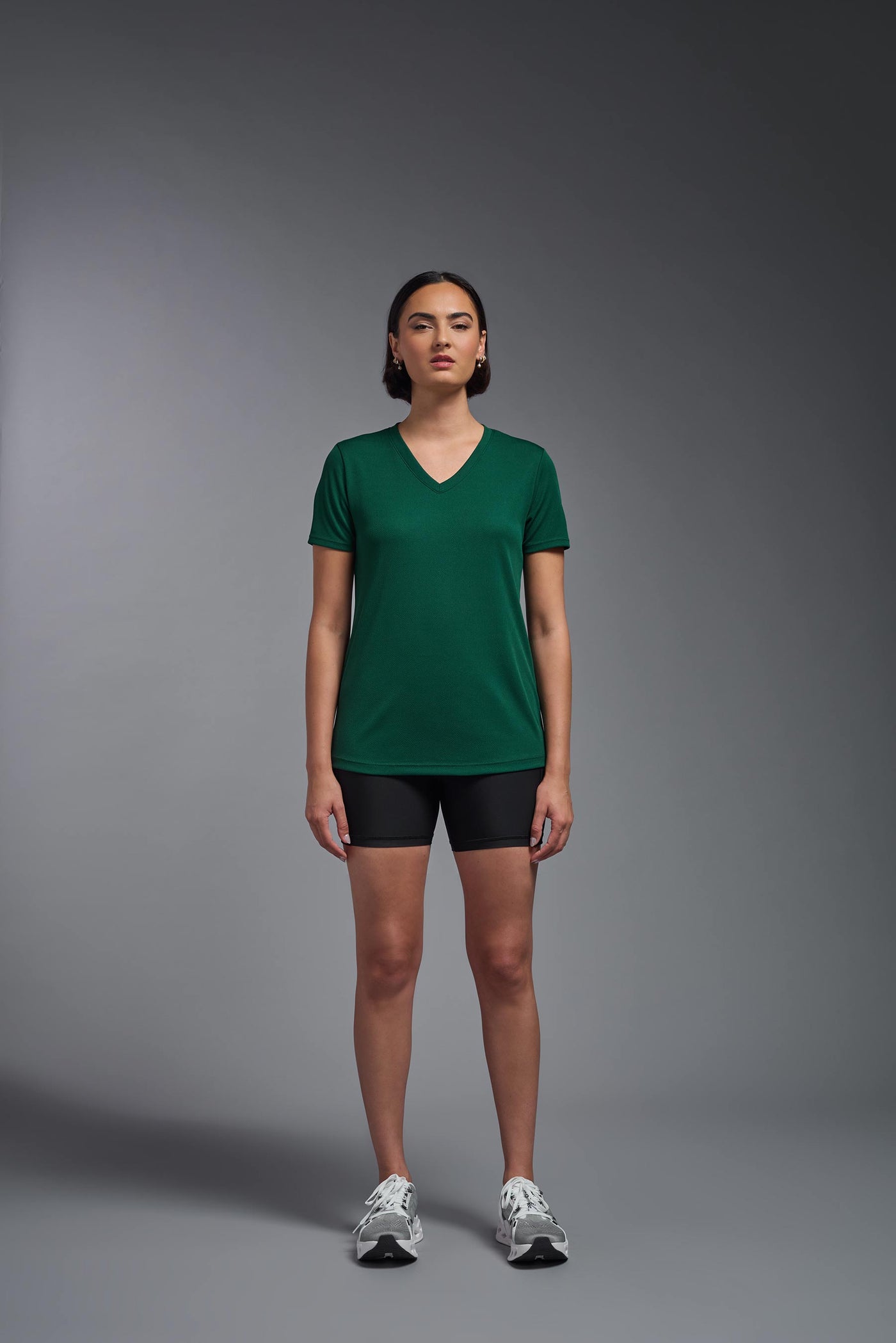 A female model with brown short hair on a gray background, wearing the Women's Oxymesh™ V-neck Tech Tee in the color forest green from a full body front view. This shirt is made in the USA and is quick-drying and moisture-wicking. #color_forest-green