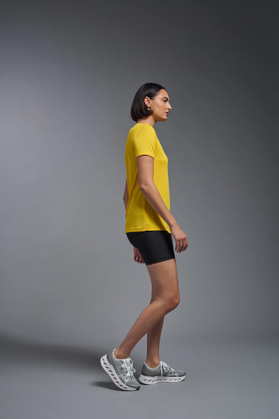 A female model with brown short hair on a gray background, wearing the Women's Oxymesh™ V-neck Tech Tee in the color gold from a full body side view. This shirt is made in the USA and is quick-drying and moisture-wicking. #color_gold
