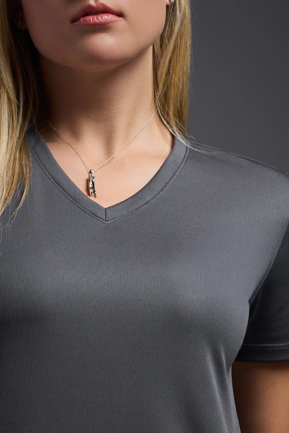 A blonde female model with long straight hair on a gray background, wearing the Women's Oxymesh™ V-neck Tech Tee in the color graphite from a closeup collar front view. This shirt is made in the USA and is quick-drying and moisture-wicking. #color_graphite-gray