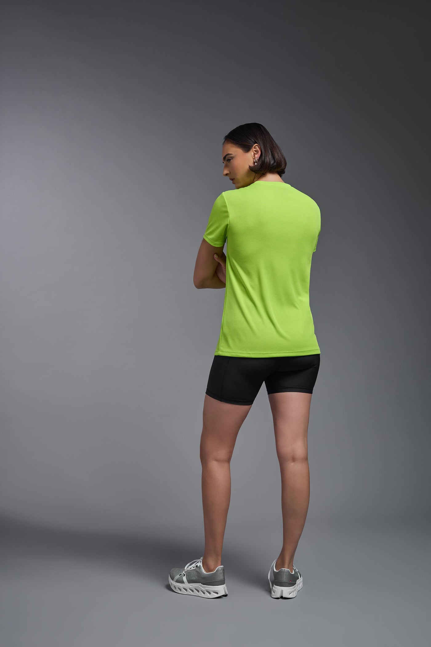 A female model with brown short hair on a gray background, wearing the Women's Oxymesh™ V-neck Tech Tee in the color key lime from a full body back view. This shirt is made in the USA and is quick-drying and moisture-wicking. #color_key-lime