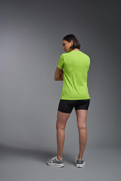 A female model with brown short hair on a gray background, wearing the Women's Oxymesh™ V-neck Tech Tee in the color key lime from a full body back view. This shirt is made in the USA and is quick-drying and moisture-wicking. #color_key-lime