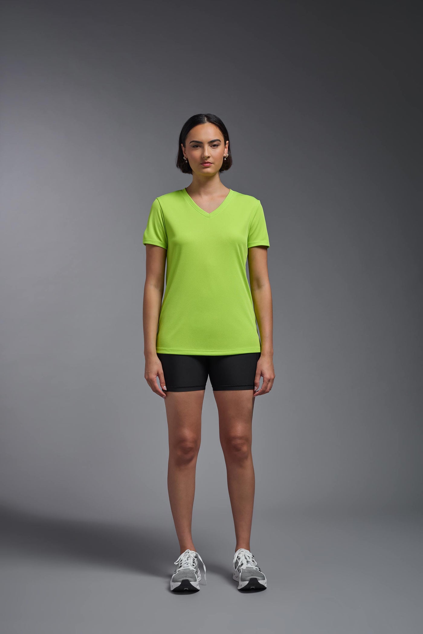 A female model with brown short hair on a gray background, wearing the Women's Oxymesh™ V-neck Tech Tee in the color key lime from a full body front view. This shirt is made in the USA and is quick-drying and moisture-wicking. #color_key-lime