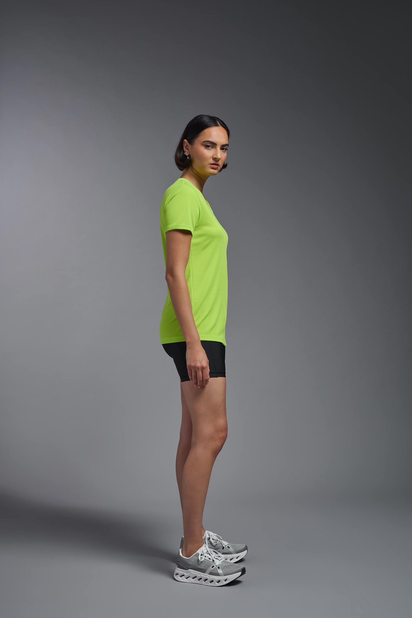 A female model with brown short hair on a gray background, wearing the Women's Oxymesh™ V-neck Tech Tee in the color key lime from a full body side view. This shirt is made in the USA and is quick-drying and moisture-wicking. #color_key-lime