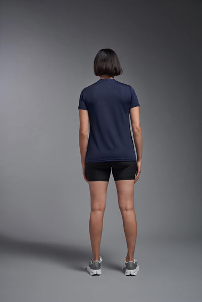 A female model with brown short hair on a gray background, wearing the Women's Oxymesh™ V-neck Tech Tee in the color navy blue from a full body back view. This shirt is made in the USA and is quick-drying and moisture-wicking. #color_navy-blue