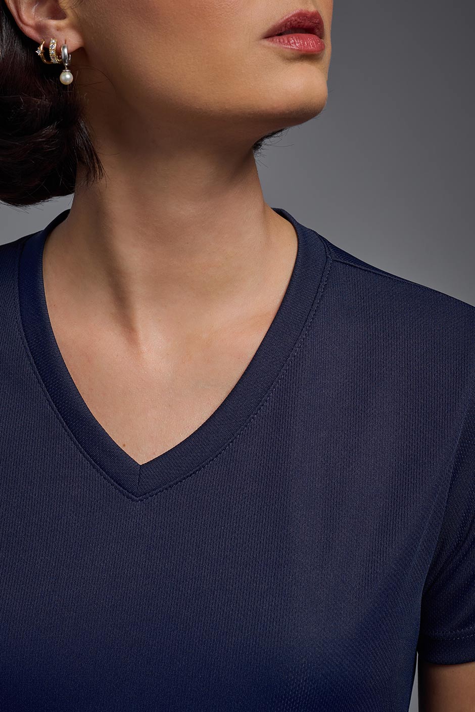 A female model with brown short hair on a gray background, wearing the Women's Oxymesh™ V-neck Tech Tee in the color navy blue from a closeup collar front view. This shirt is made in the USA and is quick-drying and moisture-wicking. #color_navy-blue