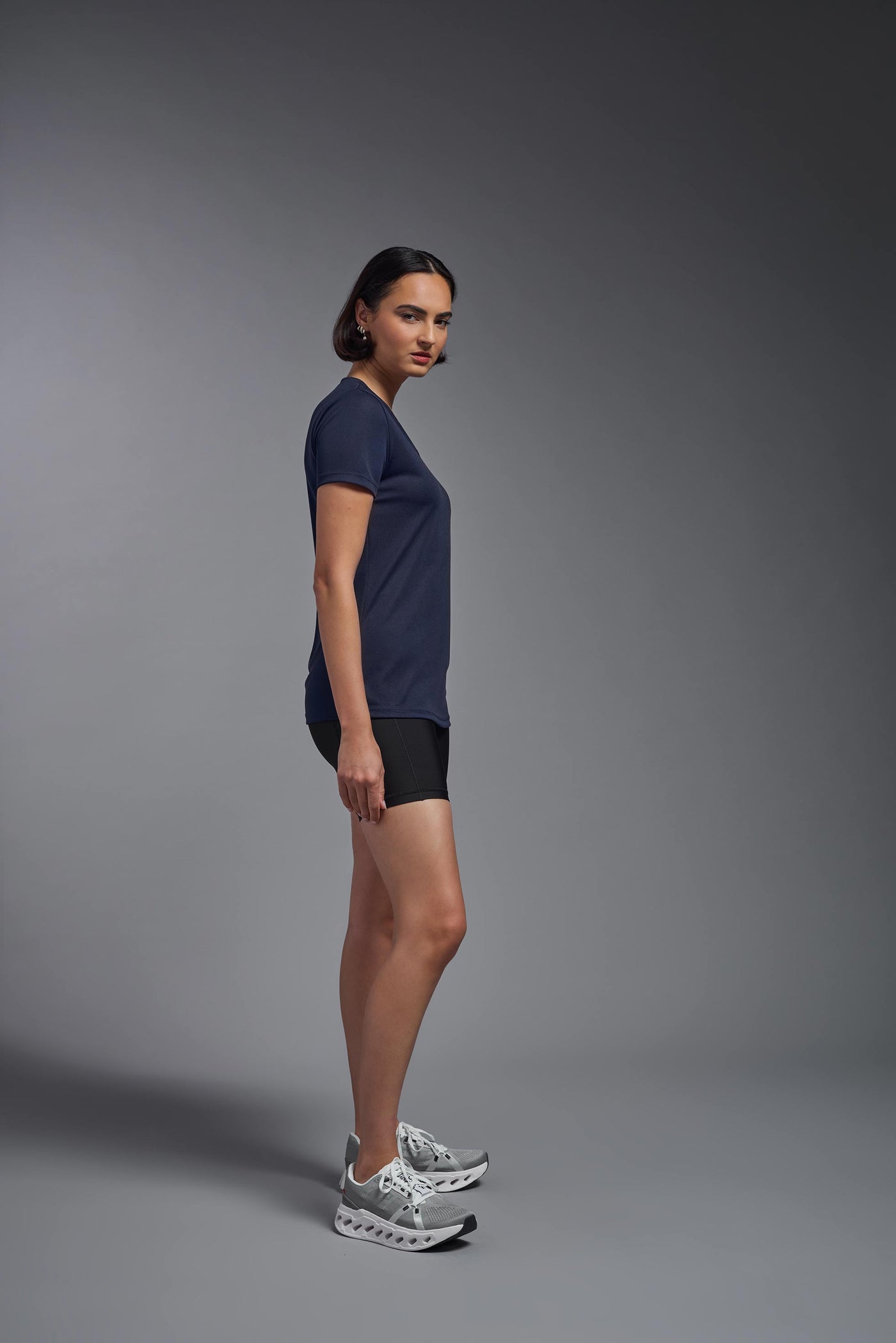 A female model with brown short hair on a gray background, wearing the Women's Oxymesh™ V-neck Tech Tee in the color navy blue from a full body side view. This shirt is made in the USA and is quick-drying and moisture-wicking. #color_navy-blue