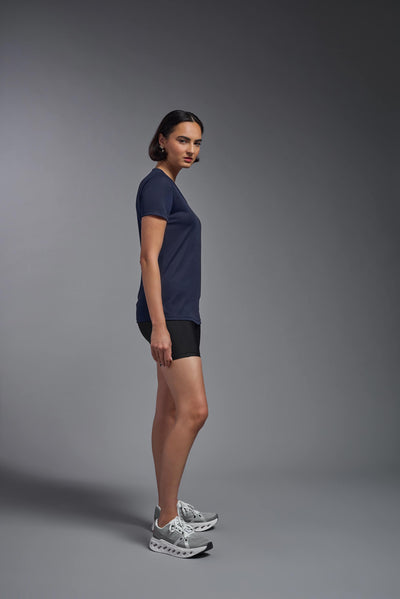 A female model with brown short hair on a gray background, wearing the Women's Oxymesh™ V-neck Tech Tee in the color navy blue from a full body side view. This shirt is made in the USA and is quick-drying and moisture-wicking. #color_navy-blue