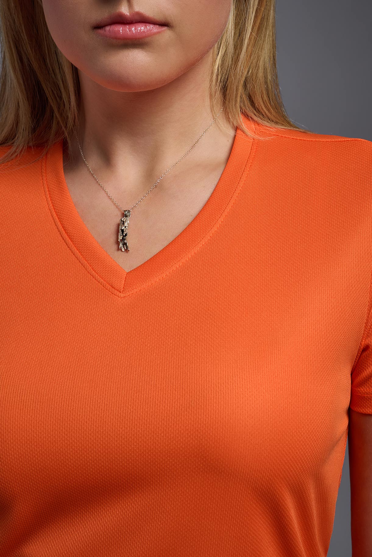 A blonde female model with long straight hair on a gray background, wearing the Women's Oxymesh™ V-neck Tech Tee in the color orange from a closeup collar front view. This shirt is made in the USA and is quick-drying and moisture-wicking. #color_orange